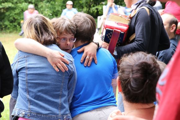 Bewohner der LebensOrte umarmen sich beim Sommerfest, ein Freund umarmt seine beiden Freunde. Akkordeonist im Hintergrund