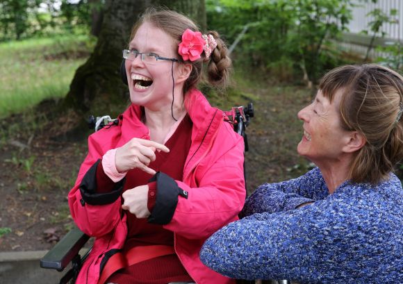 Mädchen mit Blumenschmuck im Haar lacht schallend und fröhlich, im Rollstuhl sitzend, eine lachende Betreuerin im Profil kniet neben ihr