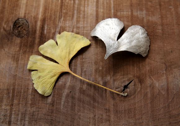 Ein Gingko-Blatt und ein hauchdünnes und zartes Kunstwerk aus unserer Designwerkstatt inform eines silbernes Gingko-Blattes auf Holzhintergrund