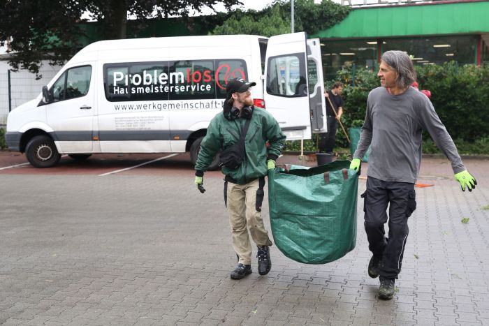 zwei Mitarbeiter von Hausmeisterei Problemlos tragen einen Sack mit Grüngut weg, im Hintergrund das Auto